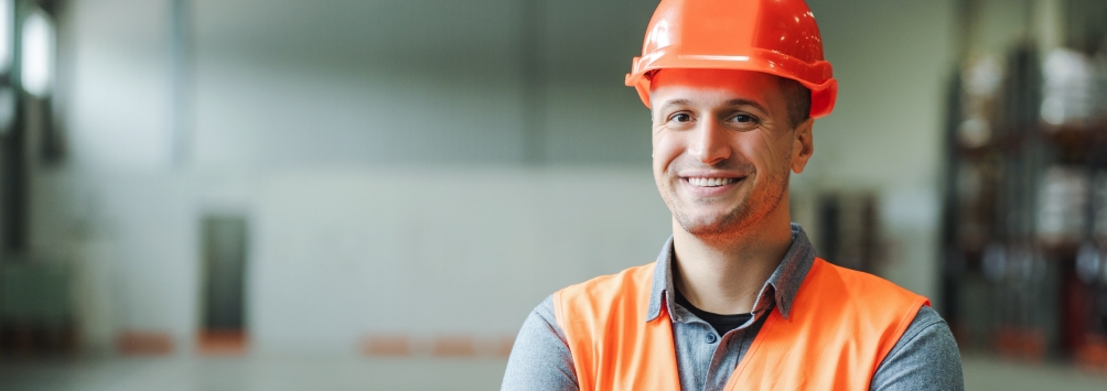 Foto de um trabalhador da industria, branco usando EPI e colete laranja sorrindo
